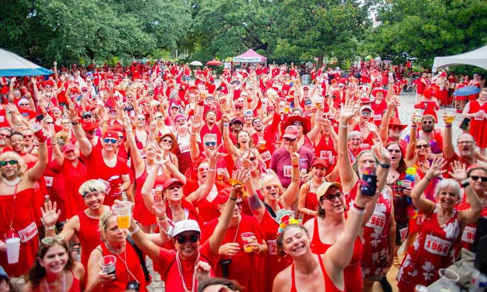 Large outdoor crowd wearing red clothing celebrating and raising hands in a forested park area during daytime.