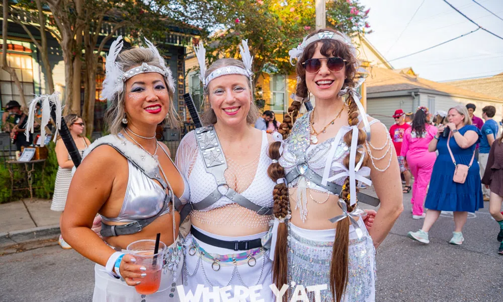 Three women in futuristic silver festival costumes posing outdoors at a sunny street event with a festive crowd.