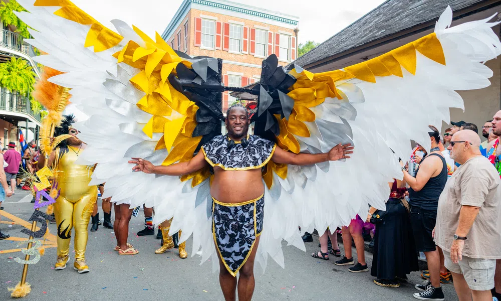Man wearing large white, gold, and black wings costume at colorful outdoor street festival parade.