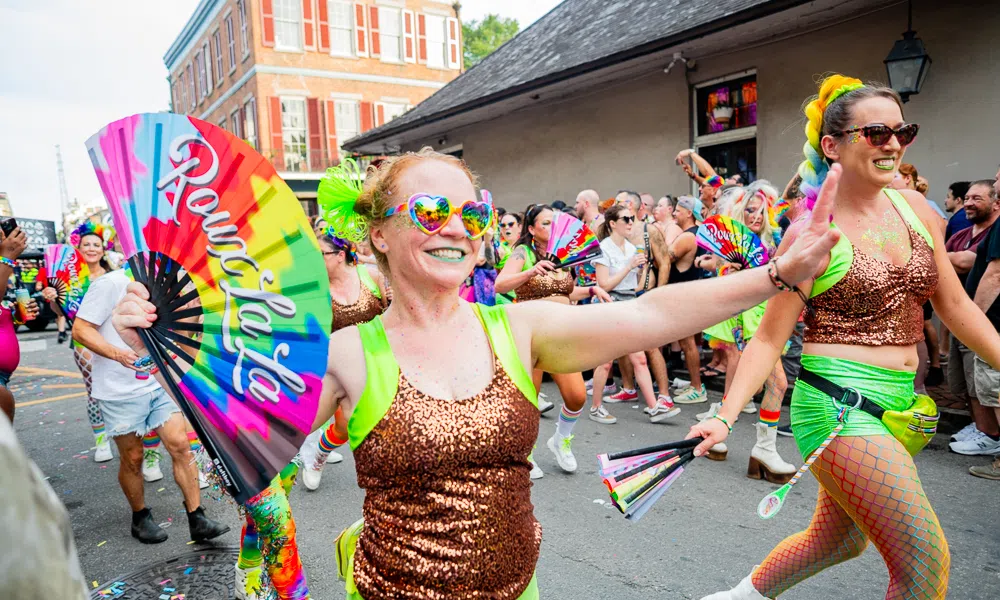 People in colorful outfits and rainbow fans celebrating during a lively street parade with smiling crowds.
