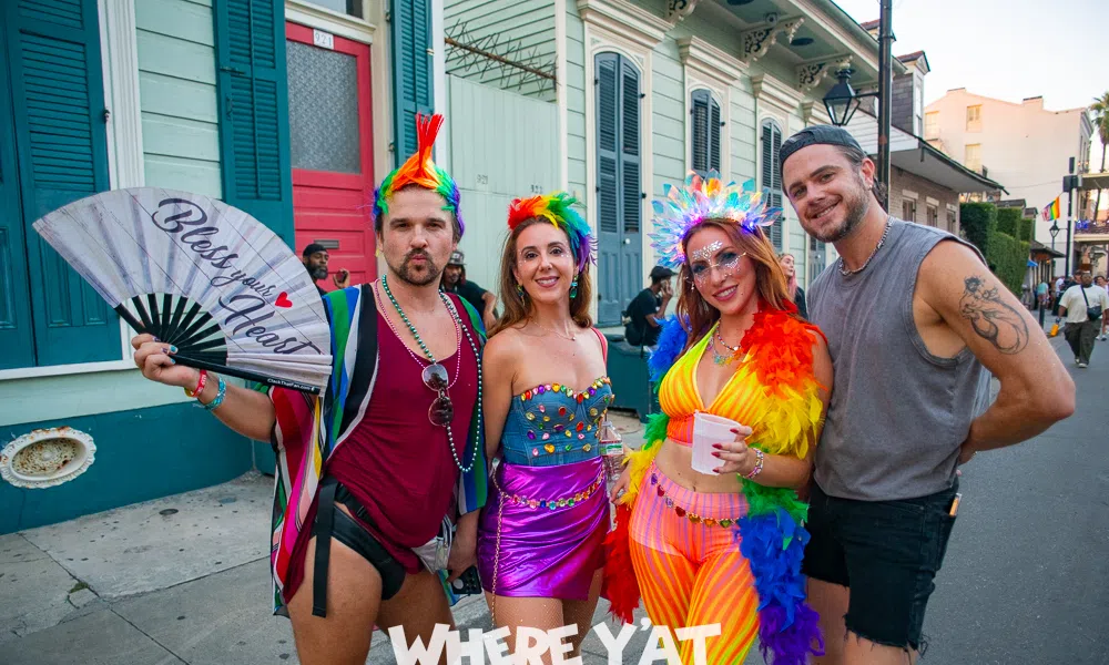 Four colorful, lively people in festive rainbow outfits posing on a New Orleans street during a celebration.
