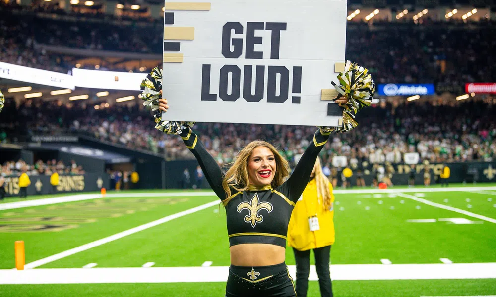Cheerleader in New Orleans Saints uniform holding a GET LOUD! sign on football field with crowd behind