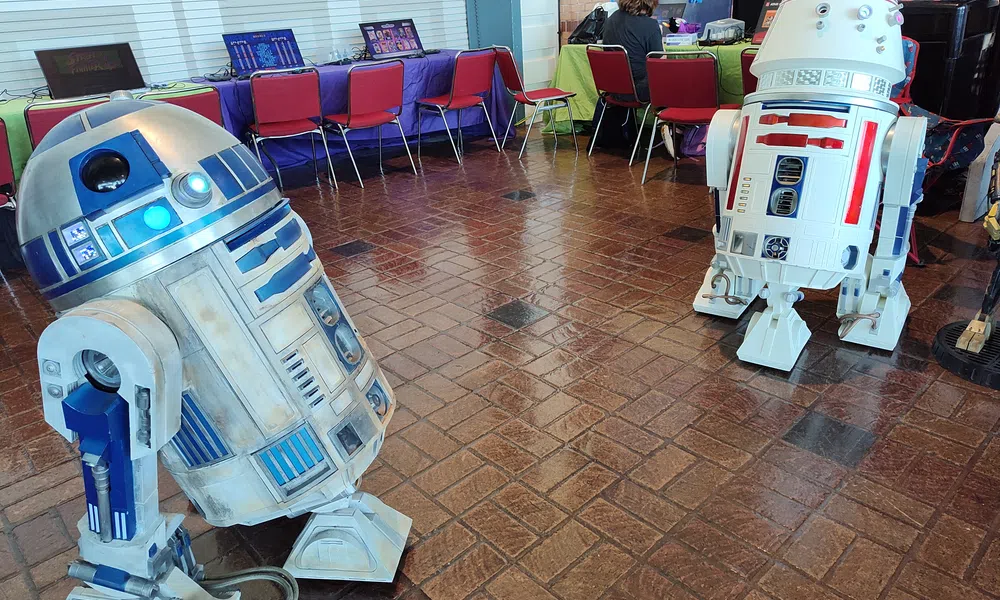 Two detailed Star Wars droids R2-D2 and an R2 variant stand on a tiled floor in a convention setting with laptops and chairs.