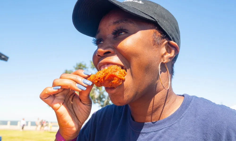 Smiling woman wearing a black cap and hoop earrings eating fried chicken outdoors on a sunny day
