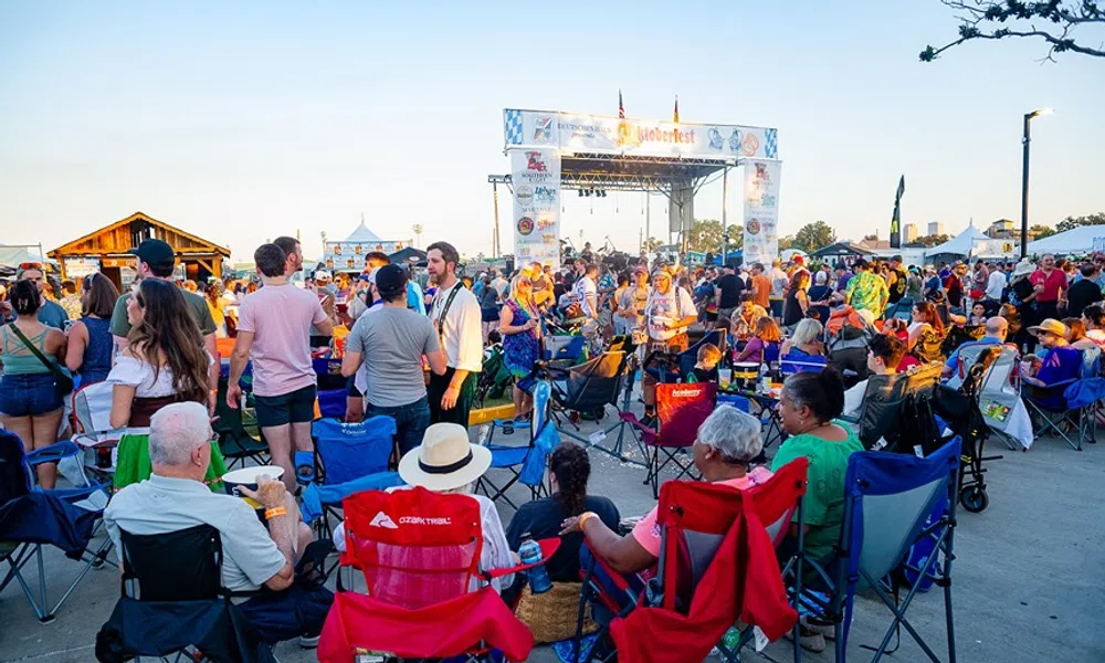 Crowd enjoying an outdoor music festival with people sitting on chairs and socializing near the stage at sunset.
