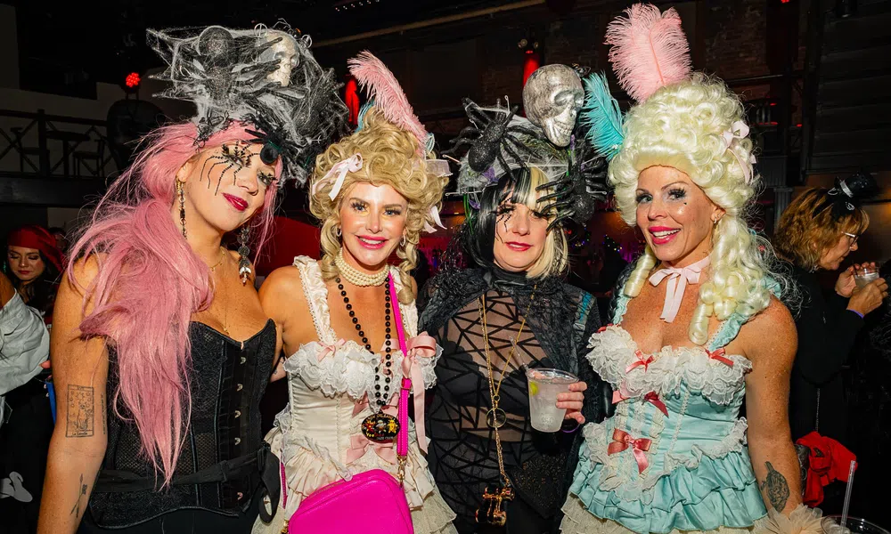 Four women in elaborate Halloween costumes with wigs and accessories at a festive party indoors.