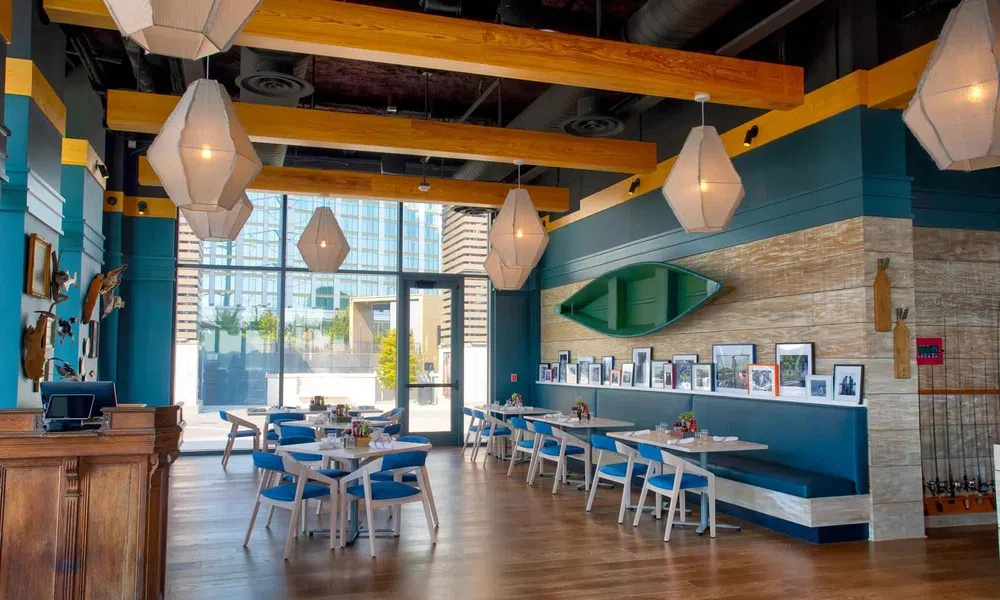 Modern dining area with wooden beams, blue chairs, pendant lights, and wall-mounted boat decor in a bright restaurant.