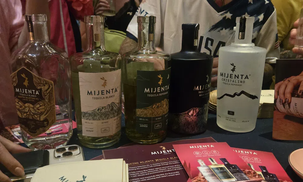 Variety of Mijenta tequila bottles displayed on table with promotional cards and people in the background.