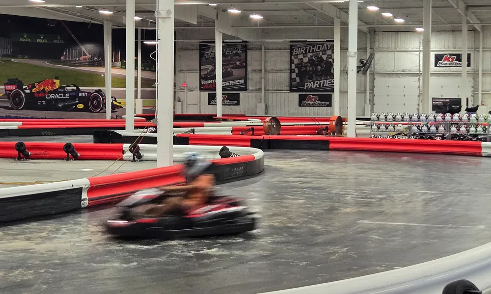 Indoor go-kart racing track with blurred driver speeding around the corner, red and white barriers, and racing posters.