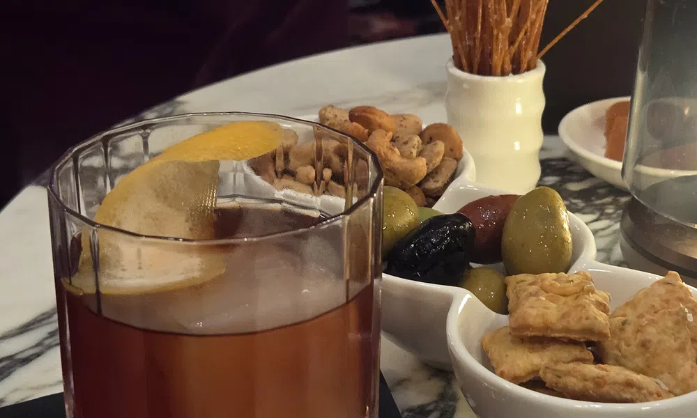 Old fashioned cocktail with lemon peel served with assorted nuts, olives, crackers, and breadsticks on marble table.