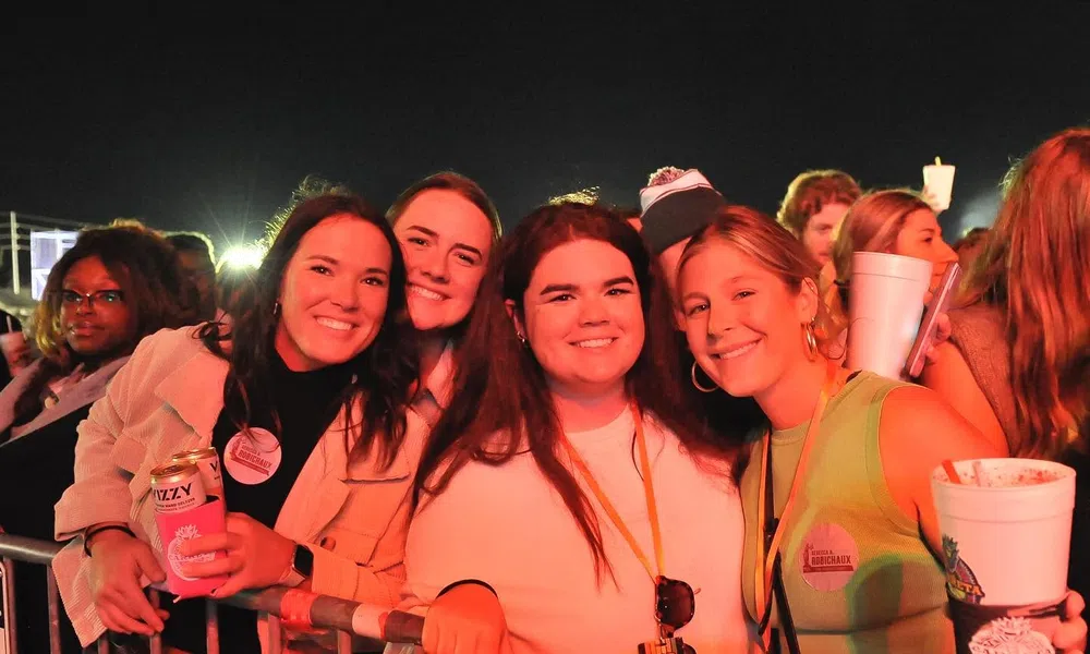 Four smiling young women posing behind a metal barrier at a nighttime outdoor event or concert, holding drinks.