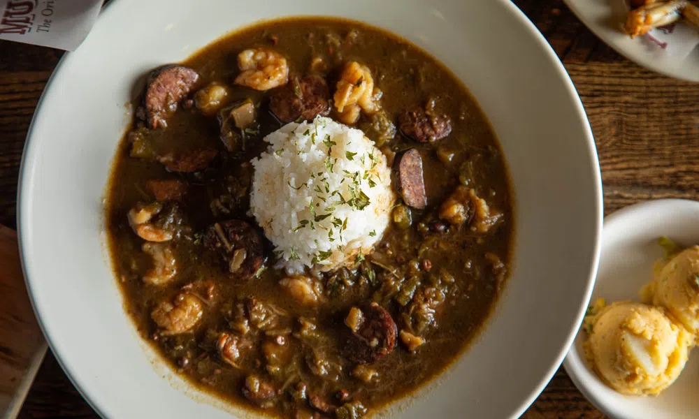 Bowl of shrimp and sausage gumbo with white rice and herbs on top, served on a wooden table with side dishes.