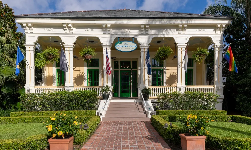 Historic country club entrance with hanging plants, flags, brick walkway, and manicured garden under blue sky