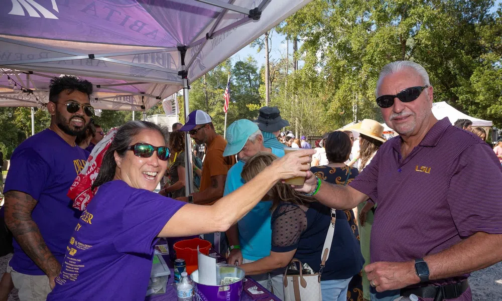 People enjoying a sunny outdoor festival, sharing drinks under a tent with vibrant purple banners.