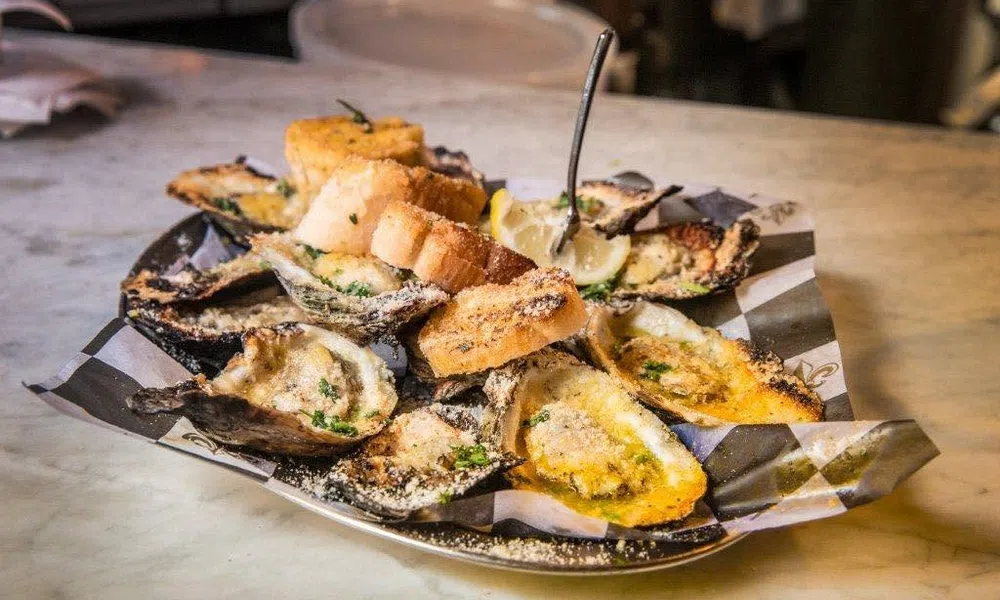 Plate of baked oysters topped with breadcrumbs, garlic, herbs, lemon wedges, and toasted bread slices on a marble table.