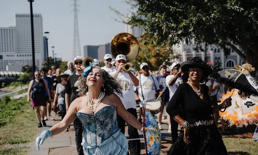 Woman in blue corset and gloves dances joyfully in front of a marching band and parade crowd on sunny day.