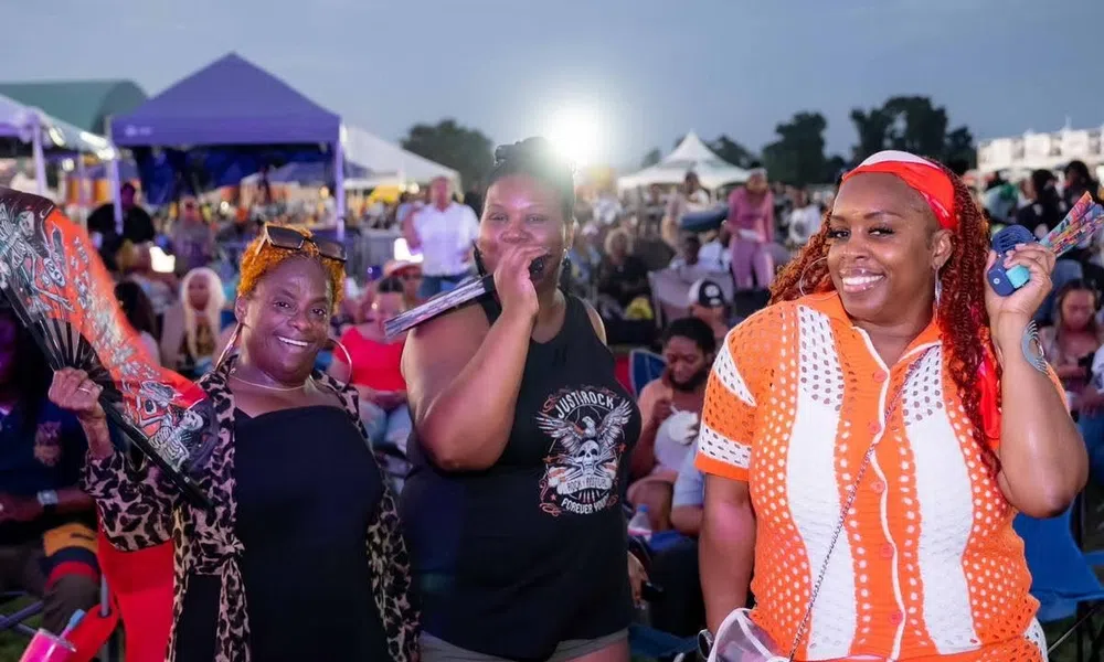 Three smiling women at an outdoor event with tents and crowd in the background during evening.