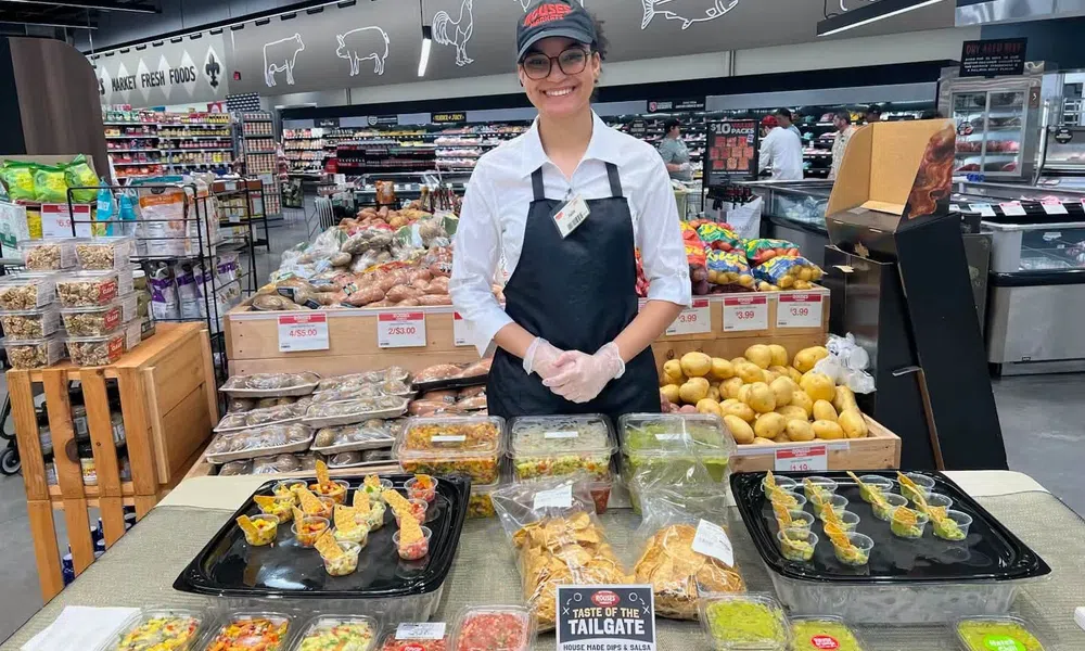 Smiling store employee presenting various homemade dips and salsas at a grocery store sampling table.