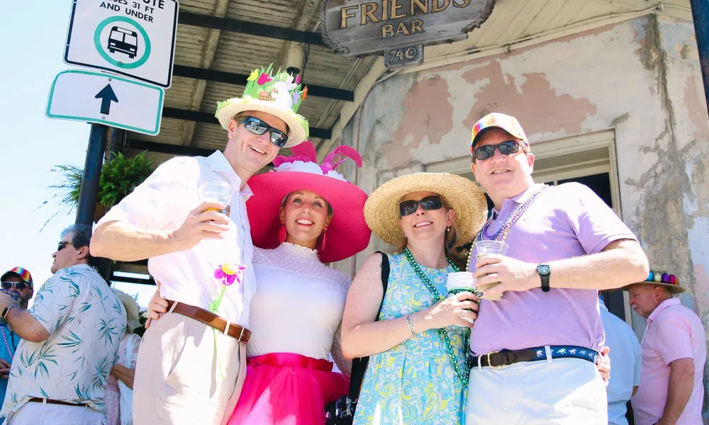 Four adults wearing colorful hats and sunglasses enjoying drinks outside Good Friends Bar on a sunny day.