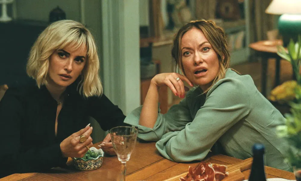 Two women with serious expressions sitting at a wooden table in a cozy, warmly lit room.