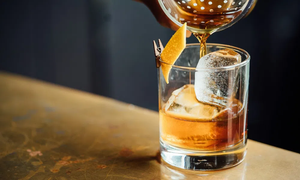 Pouring an amber cocktail over a large ice cube in a glass with a lemon peel garnish clipped to the rim