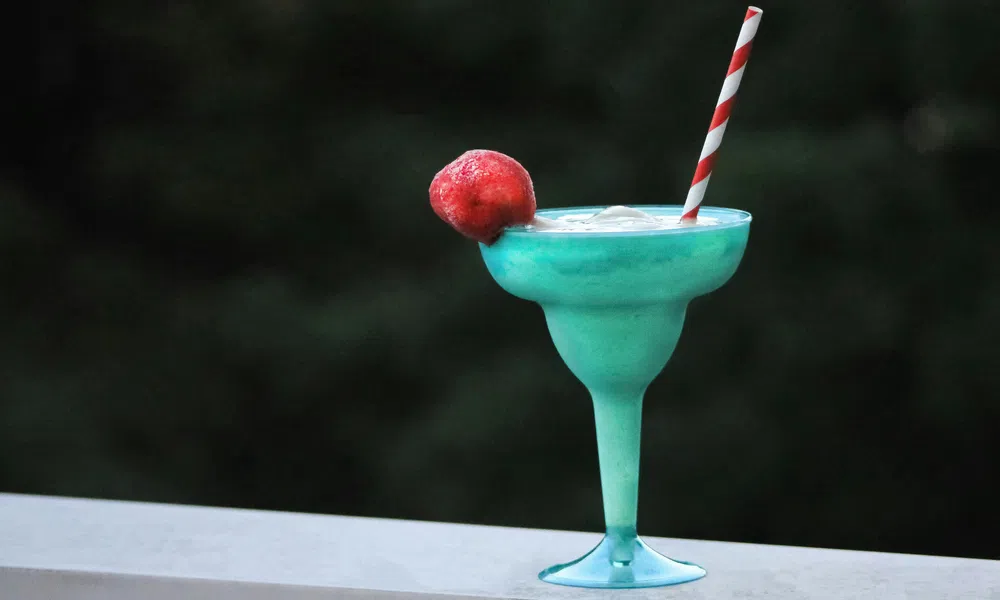 Blue tropical cocktail in a glass with ice, garnished with a red fruit and striped straw, on a white ledge.