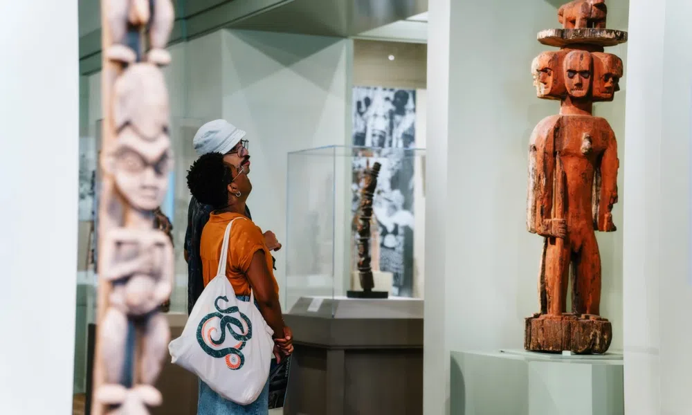 Visitors observe traditional wooden statues with carved faces at a museum exhibit focused on tribal art