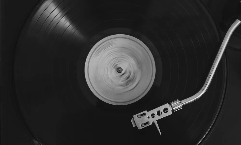 Black vinyl record spinning on a turntable with a silver tonearm in black and white photo.