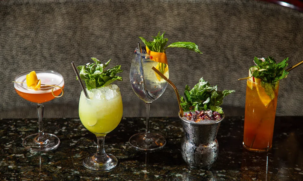 Five colorful cocktails with garnishes like mint, citrus, and berries on a dark granite bar counter.