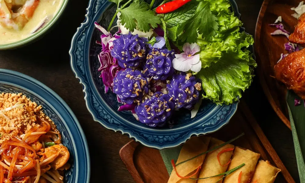 Colorful Thai dishes including purple flower-shaped dumplings, spring rolls, pad thai, and curry on ornate plates.