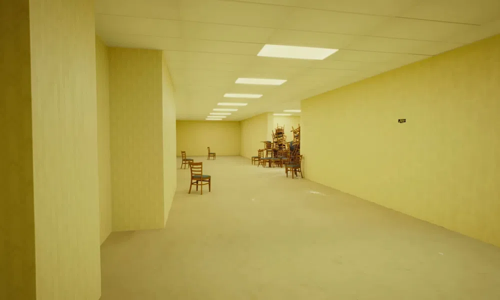 Empty beige room with chairs sparsely placed and a large stack of chairs in the corner under ceiling lights