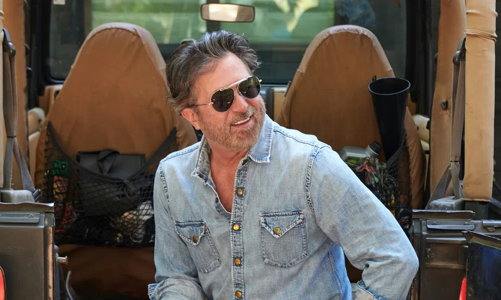 Man wearing sunglasses and denim shirt sitting in an open Jeep with tan seats smiling outdoors