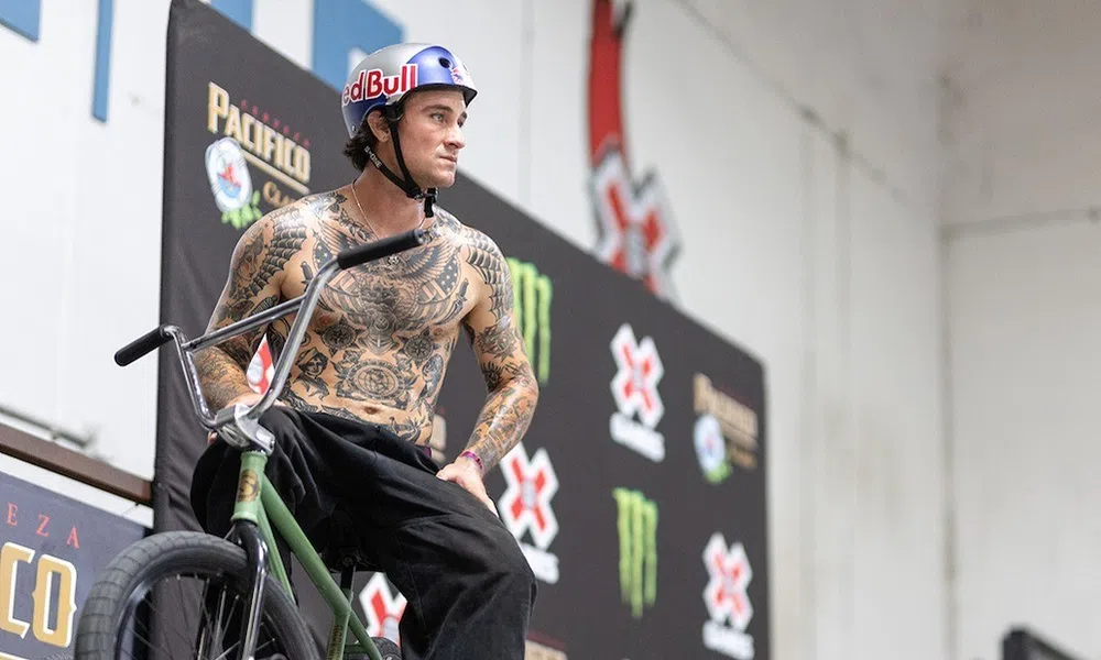 Tattooed BMX rider wearing Red Bull helmet posing with bike at indoor X Games skatepark event.