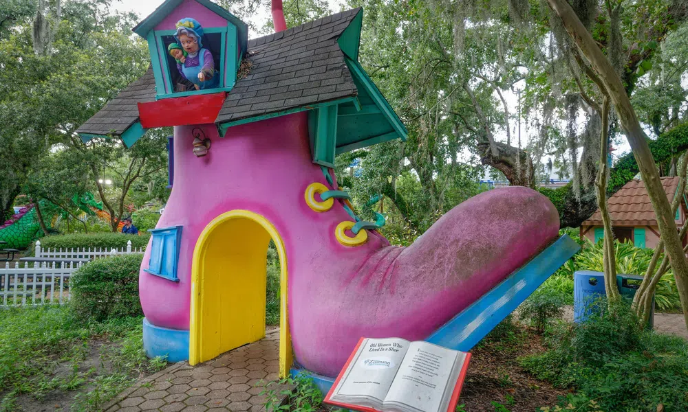 Large whimsical pink boot-shaped playhouse with blue roof and yellow door in a park setting