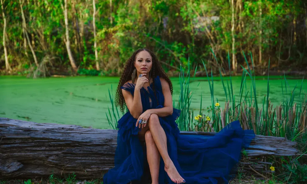 Woman in a flowing navy blue dress sitting barefoot on a log by a green pond surrounded by trees and plants.