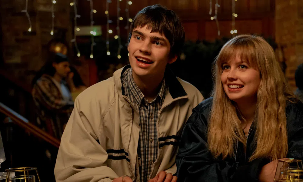 Teen boy and girl smiling and sitting together in a warmly lit indoor setting with people in the background
