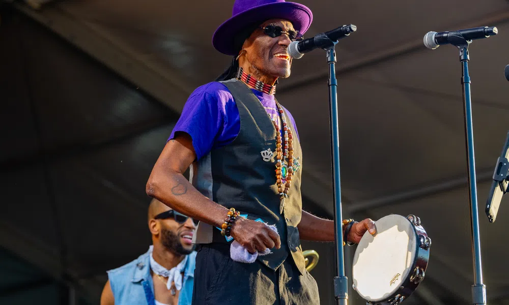 Musician in purple hat and vest singing into microphone while playing tambourine on stage at outdoor event.