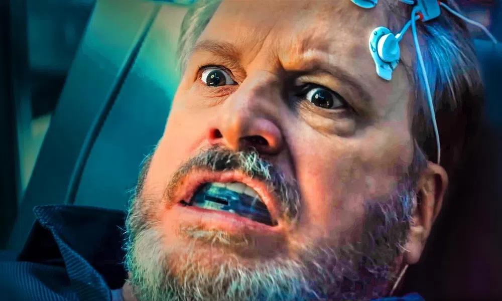 Man with electrodes attached to his head wearing a mouthguard, showing a fearful expression in a dark setting.