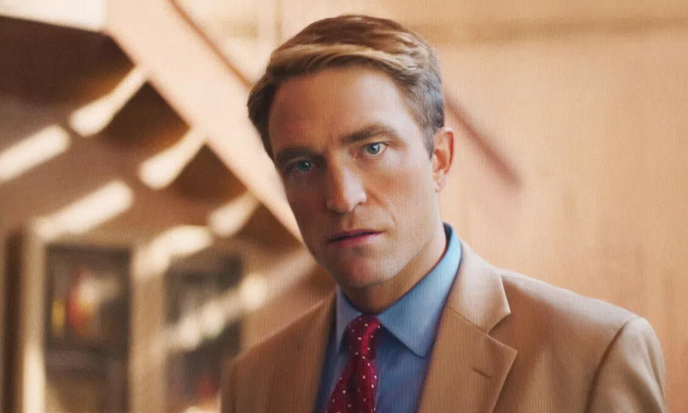 Man in a tan suit and red polka dot tie staring directly at the camera with a blurred warm background.