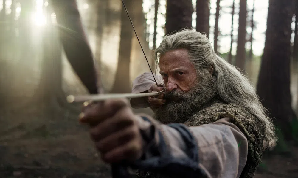Focused bearded man with long hair aiming a bow and arrow in a misty forest during daytime