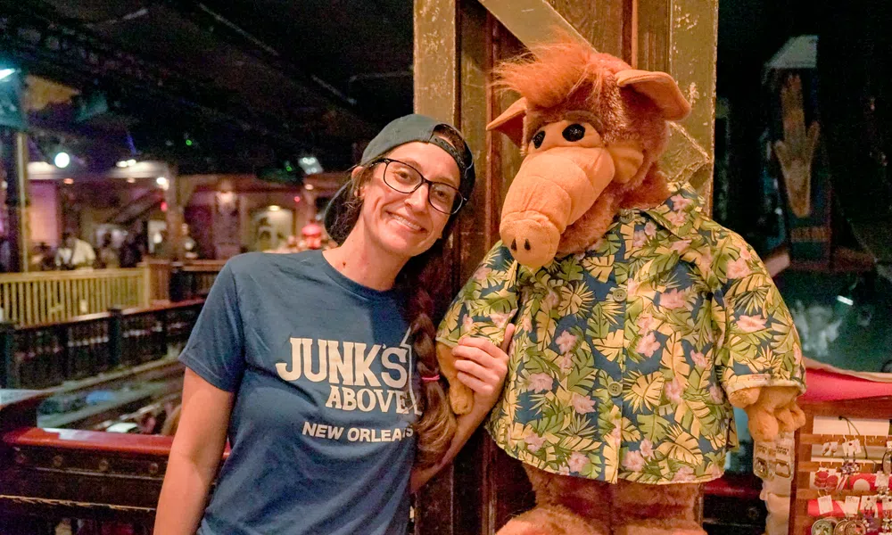 Smiling woman wearing glasses and a cap poses with ALF character doll in a tropical shirt inside a dimly lit venue.