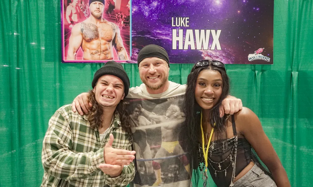 Three people smiling and posing in front of a banner featuring wrestler Luke Hawx at a convention booth.