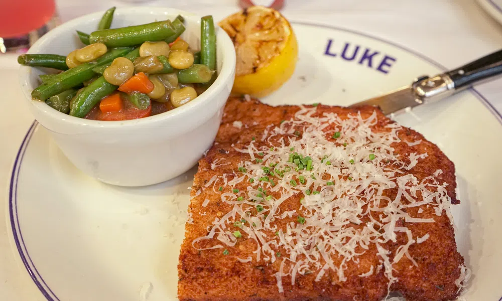 Breaded meat cutlet topped with grated cheese and herbs served with mixed vegetables and grilled lemon on a white plate.