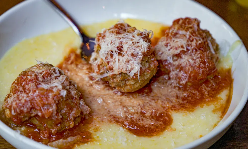 Three meatballs topped with grated cheese served in marinara sauce over creamy polenta in a white bowl.