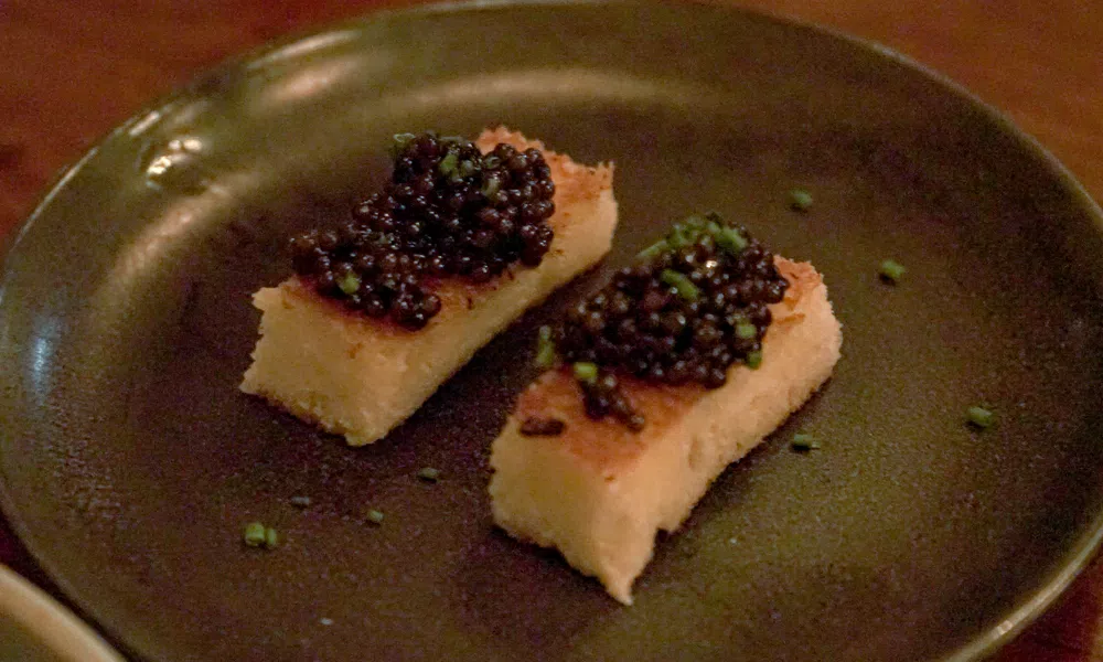 Two rectangular pieces of toasted bread topped with black caviar and garnished with chopped chives on a dark plate
