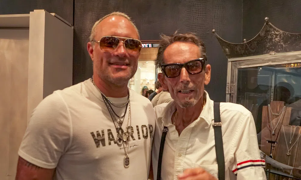 Two men wearing sunglasses and necklaces posing indoors at a jewelry store with display cases behind them