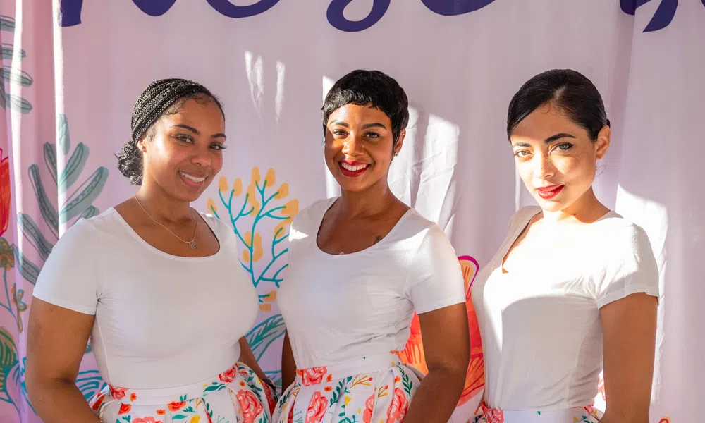Three smiling women wearing white tops and floral skirts stand in front of a colorful backdrop with plants and coral designs.