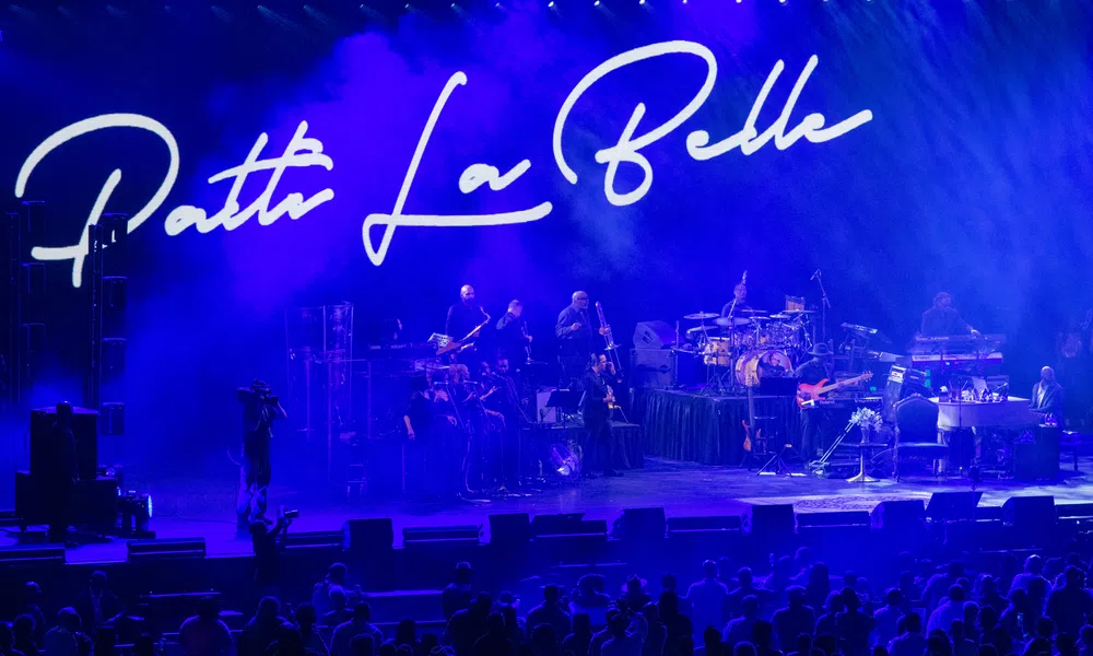 Patti LaBelle performing live on stage with band and blue lighting, audience visible in front of the stage