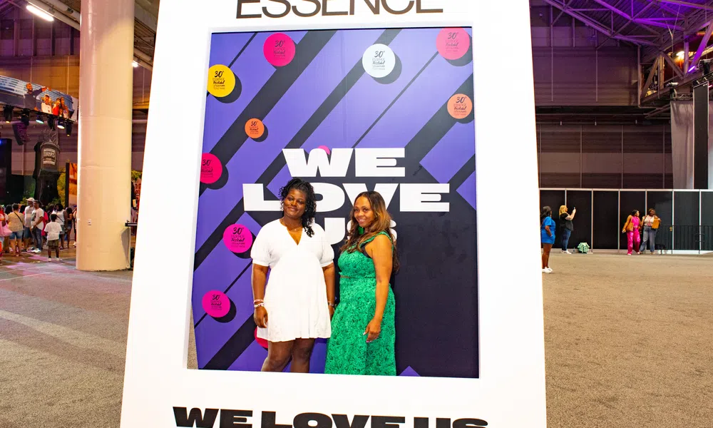 Two women posing inside a large Essence-themed photo frame with the text We Love Us at an indoor event.