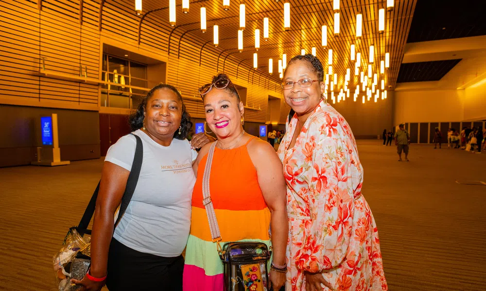 Three women smiling indoors under modern ceiling lights with wooden wall panels in a spacious venue.